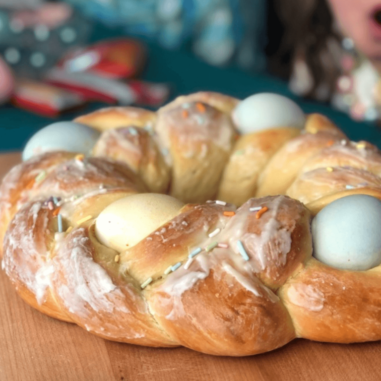 braided sourdough easter egg bread with naturally colored farm fresh eggs in the braids, topped with a glaze and naturally dyed rainbow sprinkles on a wood cutting board with kids in the background on easter morning