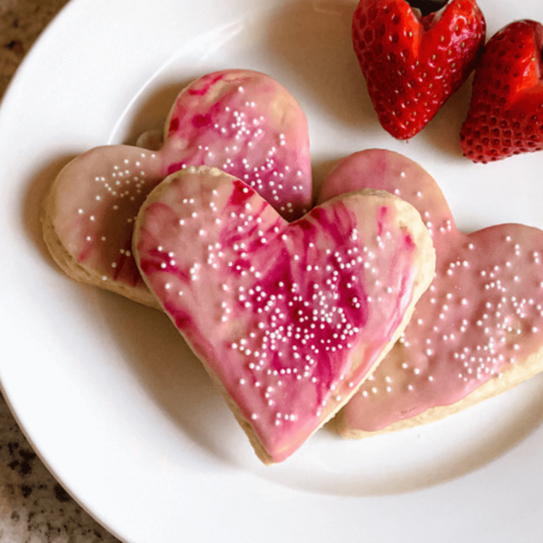 three sourdough sugar cut-out cookies shaped as hearts with marbled red and pink icing on a white plate with strawberries pared into heart shapes in upper right corner