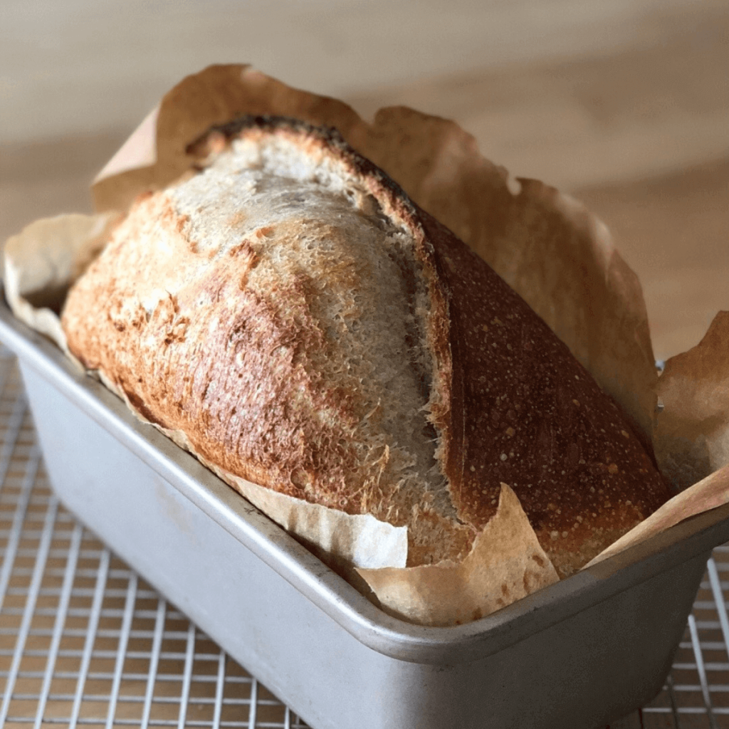 no knead sourdough sandwich loaf in a pan lined with parchment paper resting on a cooling rack for featured image of ultimate guide to no knead sourdough post