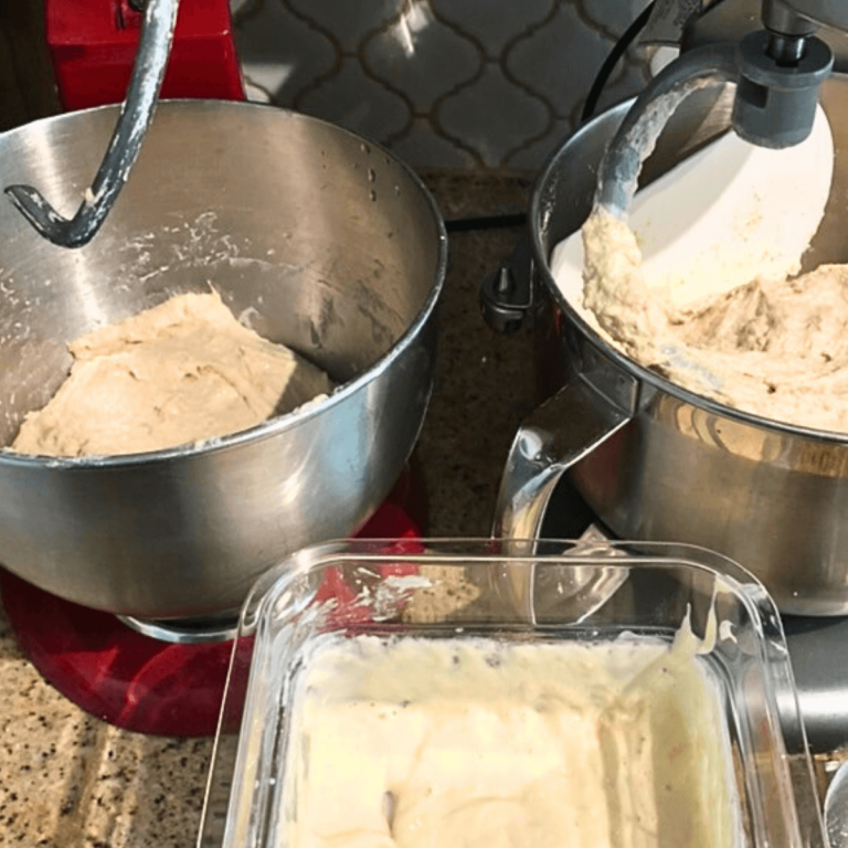 two kitchen stand mixers side by side with dough hooks attached and a batch of dough in each bowl with sourdough starter in a small cambro bin in foreground