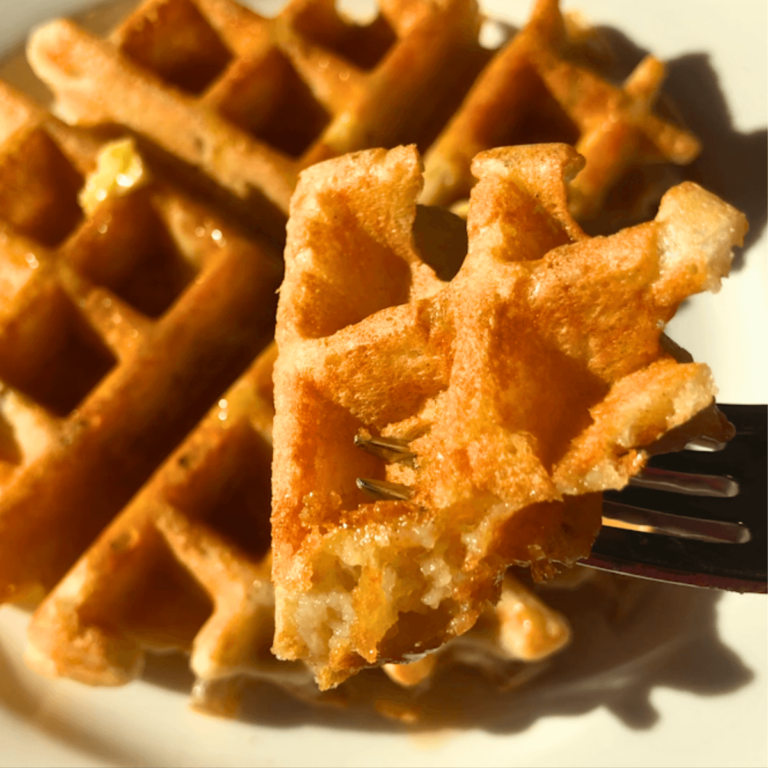 fork holding a piece of sourdough waffle covered in butter and syrup with the rest of the waffle on a white plate in the background